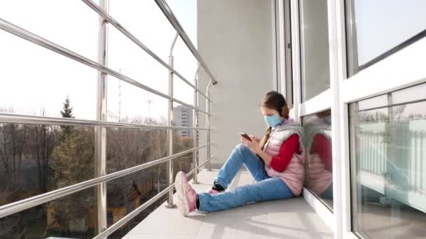 戴着面具和耳机的少女，使用智能手机，自拍，在露天阳台上。春天阳光灿烂的日子。隔离的概念，在线聊天。呆在家里。Coronavirus流行病. — 图库视频影像