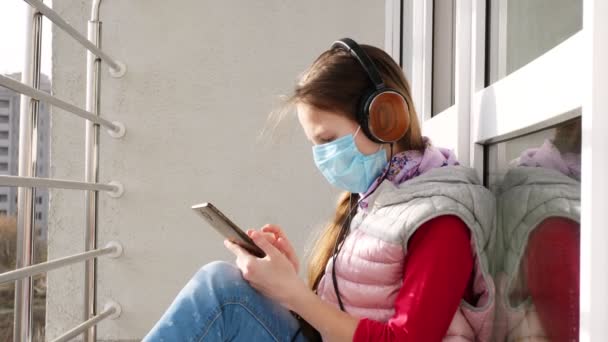 戴着面具和耳机的少女，在露天阳台上使用智能手机。春天阳光灿烂的日子。隔离的概念，在线聊天。呆在家里。Coronavirus流行病. — 图库视频影像