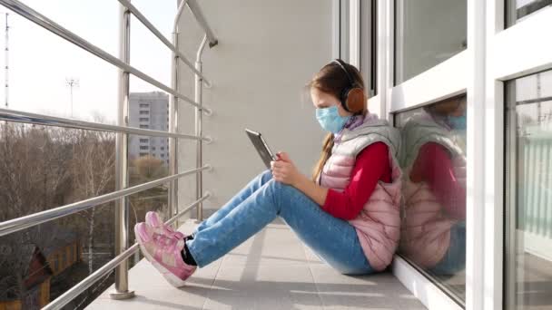 Maskeli ve kulaklıklı genç bir kız, yazı yazıyor, tablet üzerinde çalışıyor, açık balkonda çalışıyor. Bahar güneşli bir gün. Karantina sırasında uzak mesafe öğrenimi. Kal, evde çalış. Koronavirüs salgını. — Stok video