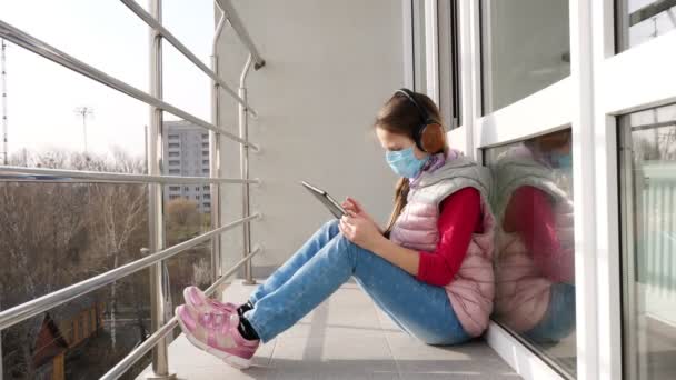 Adolescente en masque et écouteurs, dactylographiant, travaillant sur tablette, sur balcon ouvert. printemps journée ensoleillée. enseignement à distance pendant la quarantaine. Reste étudier à la maison. épidémie de coronavirus . — Video