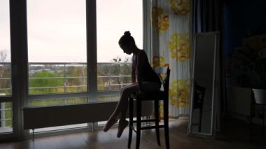 Koyu renk balerin silueti, bale mayosu giymiş kız, puanlı egzersizler yaptıktan sonra sandalyede dinleniyor. Evde bale dersleri, online dersler. büyük pencerenin yanında