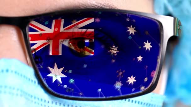 Closeup, eye, part of doctor face in medical mask, glasses, which painted in colors of Australia flag. Many viruses, germs moving on glass.State interests in vaccines, drugs invention, pathogenic — Stock Video