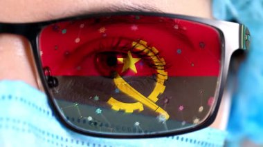 Yakın plan, göz, tıbbi maskeli bir doktor yüzü, Angola bayrağının renklerine boyanmış gözlükler. Birçok virüs, camlarda hareket eden mikroplar. Aşılara, ilaç icatlarına, patojenik virüslere devlet ilgisi.