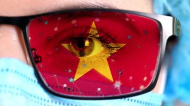 Yakın plan, göz, tıbbi maskeli bir doktor yüzü, Vietnam bayrağının renklerine boyanmış gözlükler. Birçok virüs, camlarda hareket eden mikroplar. Aşılara, uyuşturucu icatlarına, patojenik ilaçlara devletin ilgisi.
