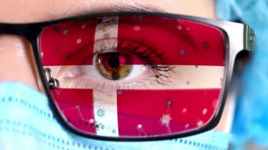 Yakın plan, göz, tıbbi maskeli doktor yüzü Danimarka bayrağının renklerine boyanmış gözlükler. Birçok virüs, camlarda hareket eden mikroplar. Aşılara, uyuşturucu icatlarına, patojenik ilaçlara devletin ilgisi.