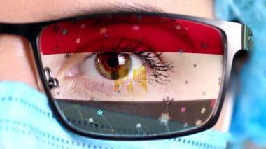 Yakın plan, göz, tıbbi maskeli doktor yüzü, gözlük, Mısır bayrağı renginde boyanmış. Birçok virüs, camlarda hareket eden mikroplar. Aşılara, ilaç icatlarına, patojenik virüslere devlet ilgisi.