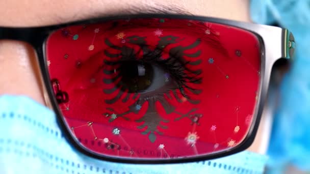 Close-up, oog, deel van de arts gezicht in medische masker, bril, die geschilderd in kleuren van Albanië vlag. Veel virussen, ziektekiemen bewegen zich op glas. Staatsbelangen in vaccins, medicijnen uitvinding, pathogene — Stockvideo