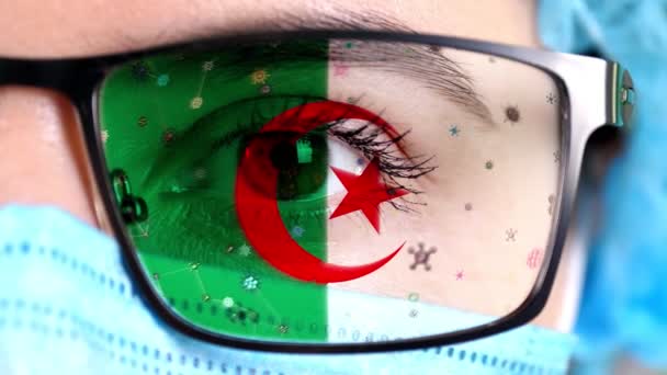 Primer plano, ojo, parte de la cara del médico en máscara médica, gafas, que pintó en colores de la bandera de Argelia. Muchos virus, gérmenes que se mueven sobre el vidrio.Intereses estatales en las vacunas, la invención de medicamentos, patógenos — Vídeo de stock