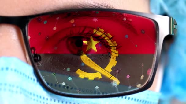 Detailní záběr, oko, část lékařského obličeje v lékařské masce, brýle, které malují v barvách angolské vlajky. Mnoho virů, bakterií pohybujících se na skle.Státní zájmy ve vakcínách, vynálezy léků, patogenní viry — Stock video