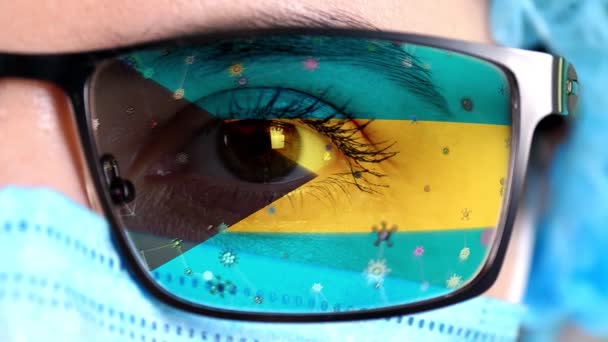 Närbild, öga, en del av läkare ansikte i medicinsk mask, glasögon, som målade i färger av Bahamas flagga. Många virus, bakterier som rör sig på glas.Statliga intressen i vacciner, droger uppfinning, patogena — Stockvideo