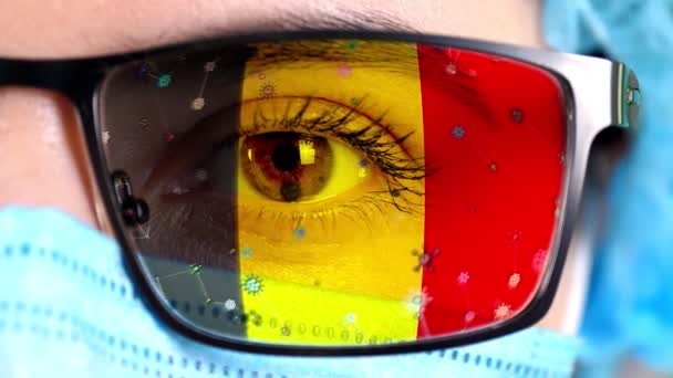 Close-up, oog, deel van het gezicht van de arts in medisch masker, bril, die geschilderd in kleuren van de Belgische vlag. Veel virussen, ziektekiemen bewegen zich op glas. Staatsbelangen in vaccins, medicijnen uitvinding, pathogene — Stockvideo