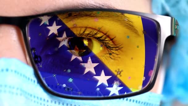 Close-up, oog, deel van de arts gezicht in medisch masker, bril, die geschilderd in de kleuren van Bosnië en Herzegovina vlag. Veel virussen, ziektekiemen bewegen zich op glas. Staatsbelangen in vaccins, drugs uitvinding — Stockvideo