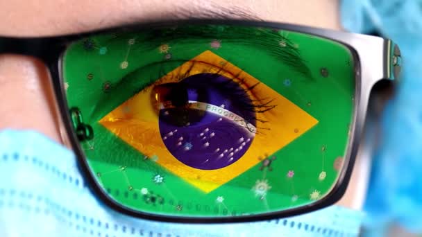 Primer plano, ojo, parte de la cara del médico en máscara médica, gafas, que pintó en colores de la bandera de Brasil. Muchos virus, gérmenes que se mueven sobre el vidrio.Intereses estatales en las vacunas, la invención de medicamentos, virus patógenos — Vídeos de Stock