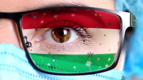 Primer plano, ojo, parte de la cara del médico en máscara médica, gafas, que pintó en colores de la bandera de Hungría. Muchos virus, gérmenes que se mueven sobre el vidrio.Intereses estatales en las vacunas, la invención de medicamentos, patógenos — Vídeo de stock