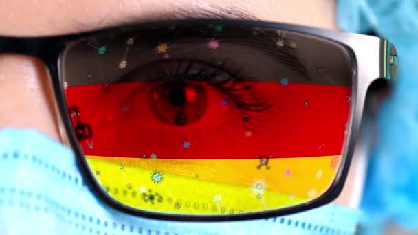 Close-up, oog, deel van de arts gezicht in medische masker, bril, die geschilderd in kleuren van Duitsland vlag. Veel virussen, ziektekiemen bewegen zich op glas. Staatsbelangen in vaccins, medicijnen uitvinding, pathogene — Stockvideo