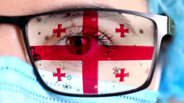 Primer plano, ojo, parte de la cara del médico en máscara médica, gafas, que pintó en colores de la bandera de Georgia. Muchos virus, gérmenes que se mueven sobre el vidrio.Intereses estatales en las vacunas, la invención de medicamentos, patógenos — Vídeos de Stock