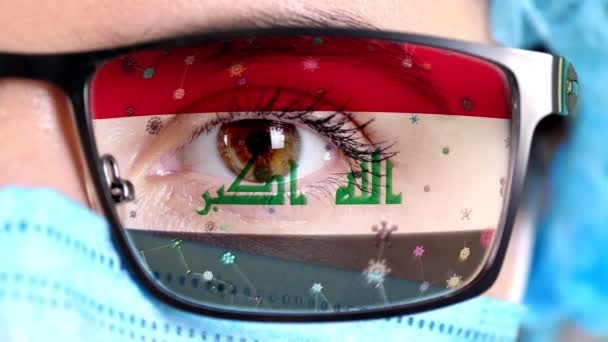 Primer plano, ojo, parte de la cara del médico en máscara médica, gafas, que pintó en colores de la bandera de Irak. Muchos virus, gérmenes que se mueven sobre el vidrio.Intereses estatales en las vacunas, la invención de medicamentos, virus patógenos — Vídeo de stock