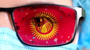 Yakın plan, göz, tıbbi maskeli doktor yüzü, Kırgızistan bayrağının renklerine boyanmış gözlükler. Birçok virüs, camlarda hareket eden mikroplar. Aşılara, uyuşturucu icatlarına, patojenik ilaçlara devletin ilgisi.