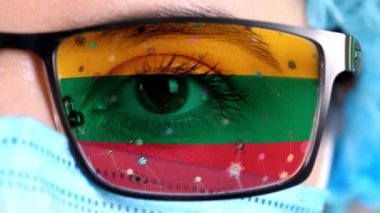 Yakın plan, göz, tıbbi maskeli doktor yüzü, gözlük, Litvanya bayrağı renginde boyanmış. Birçok virüs, camlarda hareket eden mikroplar. Aşılara, uyuşturucu icatlarına, patojenik ilaçlara devletin ilgisi.