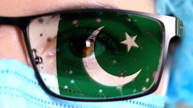 Yakın plan, göz, tıbbi maskeli bir doktor yüzü Pakistan bayrağının renklerine boyanmış gözlükler. Birçok virüs, camlarda hareket eden mikroplar. Aşılara, uyuşturucu icatlarına, patojenik ilaçlara devletin ilgisi.
