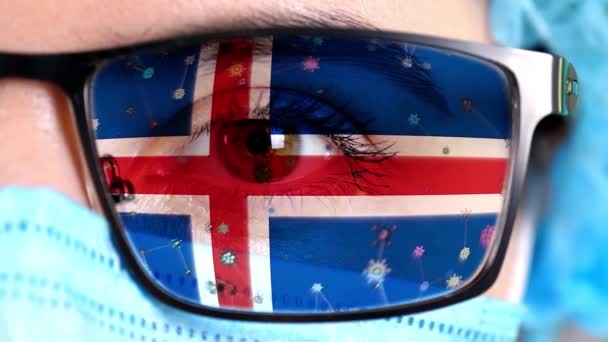 Primer plano, ojo, parte de la cara del médico en máscara médica, gafas, que pintó en colores de la bandera de Islandia. Muchos virus, gérmenes que se mueven sobre el vidrio.Intereses estatales en las vacunas, la invención de medicamentos, patógenos — Vídeo de stock