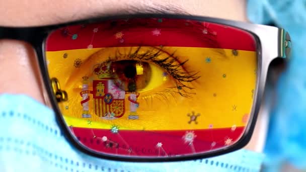 Primer plano, ojo, parte de la cara del médico en máscara médica, gafas, que pintó en colores de la bandera de España. Muchos virus, gérmenes que se mueven sobre el vidrio.Intereses estatales en las vacunas, la invención de medicamentos, virus patógenos — Vídeos de Stock