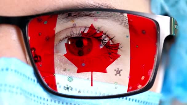 Primer plano, ojo, parte de la cara del médico en máscara médica, gafas, que pintó en colores de la bandera de Canadá. Muchos virus, gérmenes que se mueven sobre el vidrio.Intereses estatales en las vacunas, la invención de medicamentos, virus patógenos — Vídeo de stock