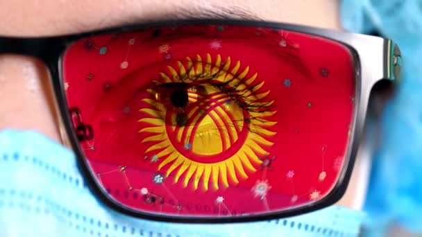 Detailní záběr, oko, část lékařského obličeje v lékařské masce, brýle, které malují v barvách kyrgyzské vlajky. Mnoho virů, bakterií pohybujících se na skle.Státní zájmy ve vakcínách, vynálezy léků, patogenní — Stock video
