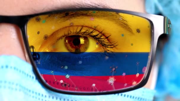 Closeup, eye, part of doctor face in medical mask, glasses, which painted in colors of Colombia flag. Many viruses, germs moving on glass.State interests in vaccines, drugs invention, pathogenic — Stock Video