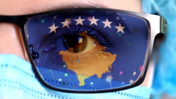 Yakın plan, göz, tıbbi maskeli bir doktor yüzü, Kosova bayrağının rengine boyanmış gözlükler. Birçok virüs, camlarda hareket eden mikroplar. Aşılara, ilaç icatlarına, patojenik virüslere devlet ilgisi. — Stok video