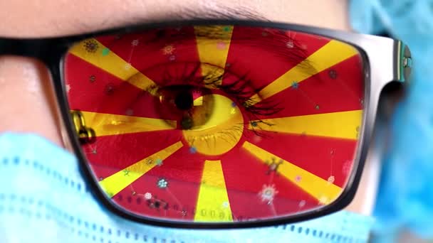 Närbild, öga, en del av läkare ansikte i medicinsk mask, glasögon, som målade i färger Makedonien flagga. Många virus, bakterier som rör sig på glas.Statliga intressen i vacciner, droger uppfinning, patogena — Stockvideo
