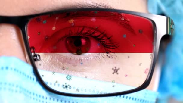 Крупный план, глаз, часть лица врача в медицинской маске, очки, которые окрашены в цвета флага Монако. Многие вирусы, микробы движутся по стеклу. Интересы государства в вакцинах, лекарственных изобретениях, патогенных вирусах — стоковое видео