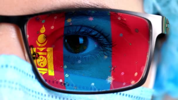 Primer plano, ojo, parte de la cara del médico en máscara médica, gafas, que pintó en colores de la bandera de Mongolia. Muchos virus, gérmenes que se mueven sobre el vidrio.Intereses estatales en las vacunas, la invención de medicamentos, patógenos — Vídeo de stock