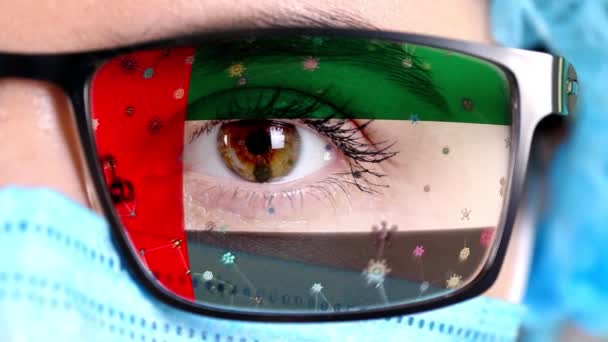Närbild, öga, en del av läkare ansikte i medicinsk mask, glasögon, som målade i färger av UAE flagga. Många virus, bakterier som rör sig på glas.Statliga intressen i vacciner, läkemedelsuppfinningar, patogena virus — Stockvideo