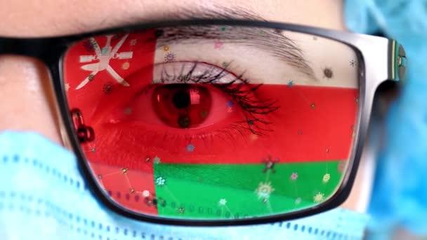 Primer plano, ojo, parte de la cara del médico en máscara médica, gafas, que pintó en colores de la bandera de Omán. Muchos virus, gérmenes que se mueven sobre el vidrio.Intereses estatales en las vacunas, la invención de medicamentos, virus patógenos — Vídeo de stock