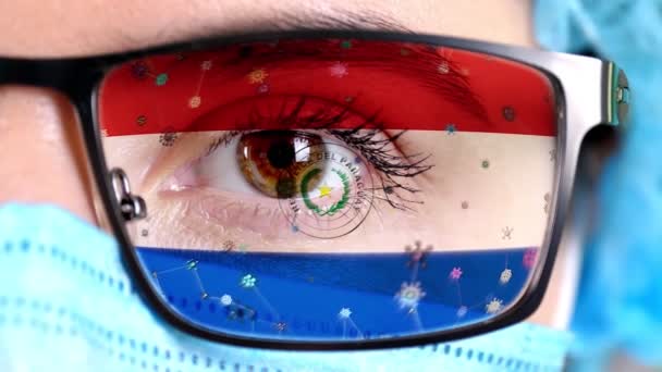 Closeup, eye, part of doctor face in medical mask, glasses, which painted in colors of Paraguay flag. Many viruses, germs moving on glass.State interests in vaccines, drugs invention, pathogenic — Stock Video