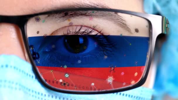 Primer plano, ojo, parte de la cara del médico en máscara médica, gafas, que pintó en colores de la bandera de Rusia. Muchos virus, gérmenes que se mueven sobre el vidrio.Intereses estatales en las vacunas, la invención de medicamentos, virus patógenos — Vídeo de stock