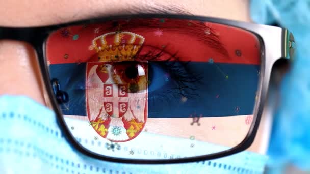 Närbild, öga, en del av läkare ansikte i medicinsk mask, glasögon, som målade i färger i Serbien flagga. Många virus, bakterier som rör sig på glas.Statliga intressen i vacciner, läkemedelsuppfinningar, patogena virus — Stockvideo