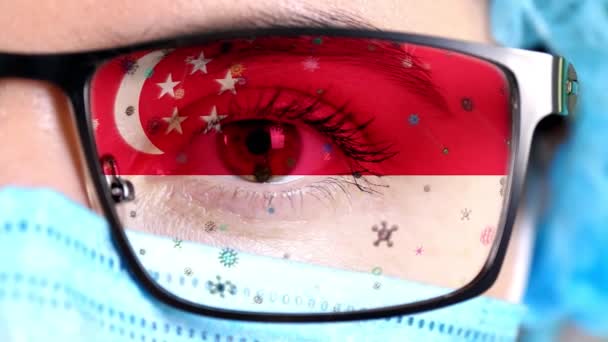 Close-up, oog, deel van de arts gezicht in medische masker, bril, die geschilderd in de kleuren van Singapore vlag. Veel virussen, ziektekiemen bewegen zich op glas. Staatsbelangen in vaccins, medicijnen uitvinding, pathogene — Stockvideo