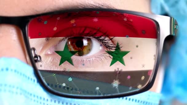 Zbliżenie, oko, część twarzy lekarza w masce medycznej, okulary, które malowane w kolorach flagi Syrii. Wiele wirusów, zarazków przemieszczających się po szkłach.Zainteresowania państwa szczepionkami, wynalazkami leków, wirusami chorobotwórczymi — Wideo stockowe