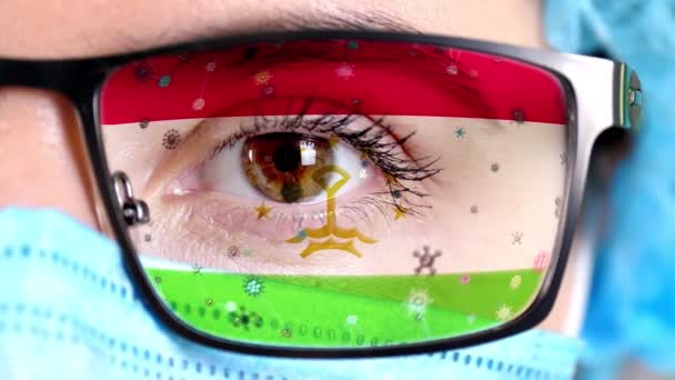 Primer plano, ojo, parte de la cara del médico en máscara médica, gafas, que pintaron en colores de la bandera de Tayikistán. Muchos virus, gérmenes que se mueven sobre el vidrio.Intereses estatales en las vacunas, la invención de medicamentos, patógenos — Vídeo de stock