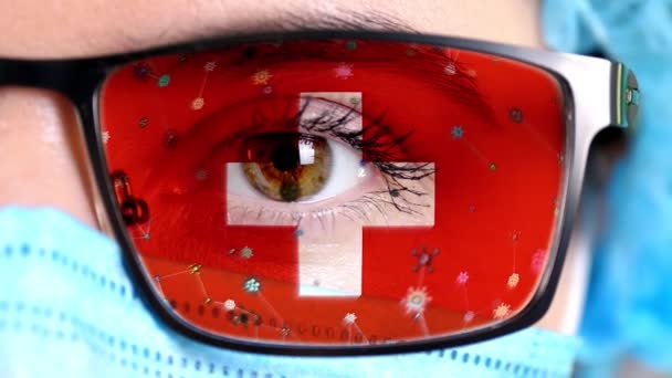 Close-up, oog, deel van de arts gezicht in medische masker, bril, die geschilderd in de kleuren van Zwitserland vlag. Veel virussen, ziektekiemen bewegen zich op glas. Staatsbelangen in vaccins, medicijnen uitvinding, pathogene — Stockvideo