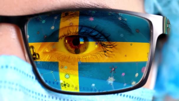 Крупный план, глаз, часть лица врача в медицинской маске, очки, которые окрашены в цвета шведского флага. Многие вирусы, микробы движутся по стеклу. Интересы государства в вакцинах, лекарственных изобретениях, патогенных вирусах — стоковое видео