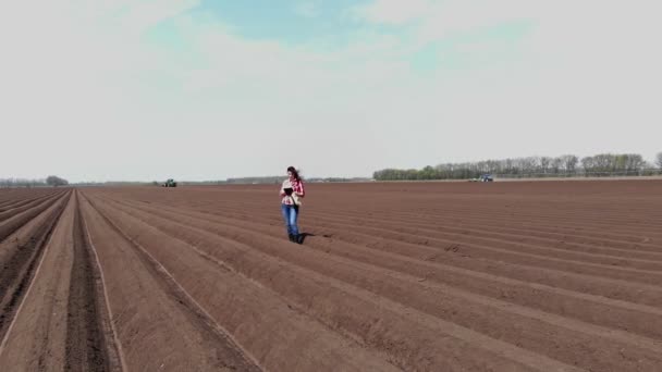 Aero. Kobieta rolnik, rolnik jest na polu ze specjalnymi rzędami gleby. Testuje, używając tabletu, jakość zautomatyzowanego sadzenia ziemniaków przez ciągniki. Nowoczesne rolnictwo, rolnictwo. wiosenny słoneczny dzień. — Wideo stockowe