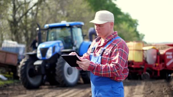 Retrato de agricultor ou agrônomo, em camisa xadrez vermelho, está digitando em smth tablet, contra fundo de tratores, máquinas agrícolas. agricultura moderna, agricultura. dia ensolarado quente — Vídeo de Stock