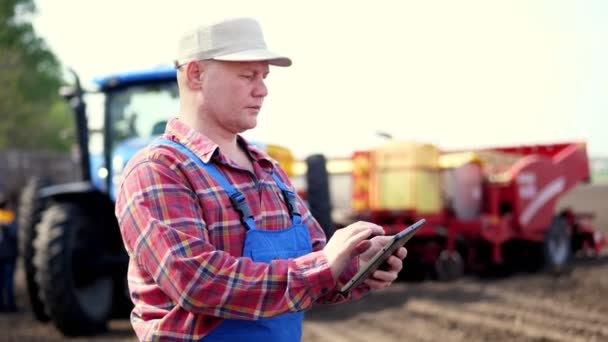 Retrato de agricultor o agrónomo, en camisa a cuadros rojo, está escribiendo en la tableta smth, sobre el fondo de tractores, maquinaria agrícola. agricultura moderna, agricultura. cálido día soleado — Vídeo de stock