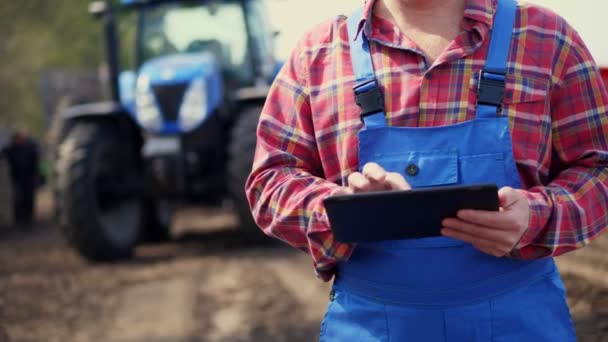 Крупним планом руки. Фермер, в червоній сорочці, друкує таблетки на тлі тракторів, сільськогосподарської техніки. сучасне сільське господарство, сільське господарство. теплий сонячний день — стокове відео
