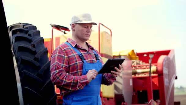 Retrato de agricultor ou agrônomo, em camisa xadrez vermelho, está digitando em smth tablet, contra fundo de tratores, máquinas agrícolas. agricultura moderna, agricultura. dia ensolarado quente — Vídeo de Stock