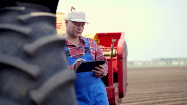 農民や農学者の肖像画は、赤い再生シャツで、トラクター、農業機械の背景を背景に、タブレットのスムーズに入力されています。農業の近代化です。暖かい晴れた日 — ストック動画