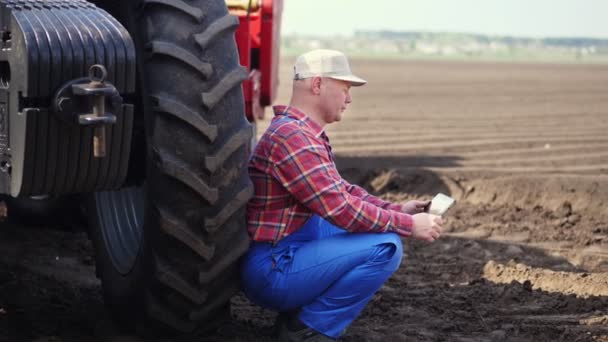 Bauer, in rotem Karohemd, tippt auf Tabletts, sitzt neben dem großen Rad des Traktors. Landmaschinen. moderne Landwirtschaft, Landwirtschaft. warmer, sonniger Tag — Stockvideo